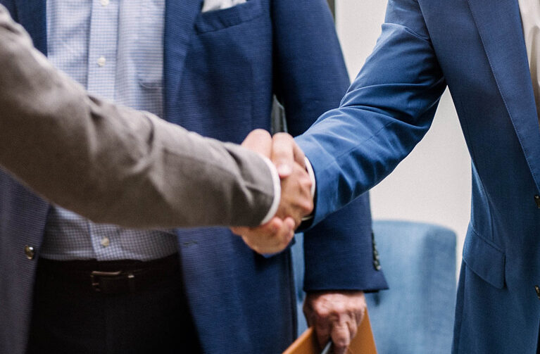 A close-up view focuses on the handshake between two men in business suits. The man on the left wears a gray jacket, and the man on the right wears a blue jacket.