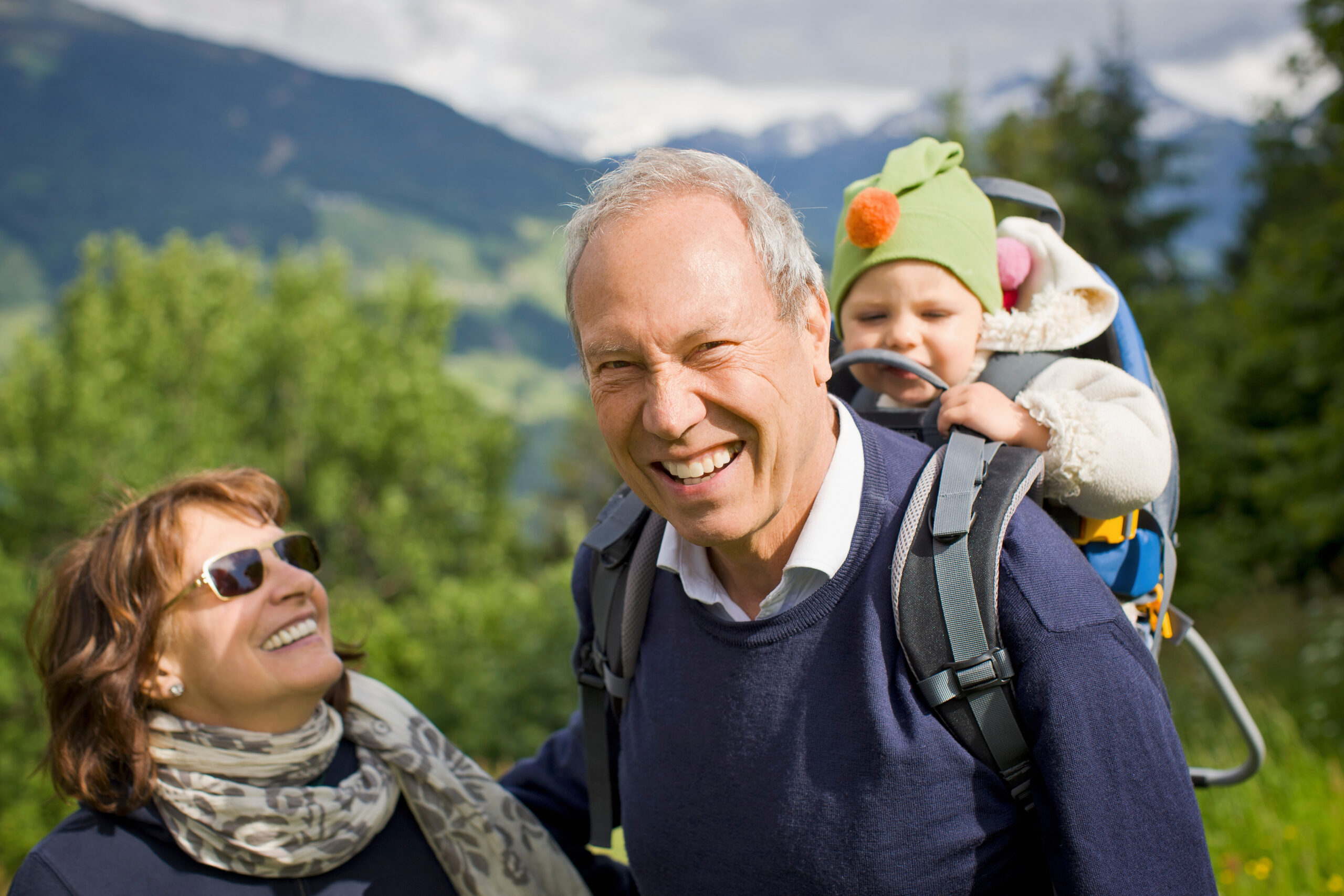 A smiling senior man, carrying a small child in a backpack carrier, stands beside a senior woman wearing sunglasses. The woman smiles and looks up at the man. They are outdoors in a grassy, mountainous setting on a sunny day.