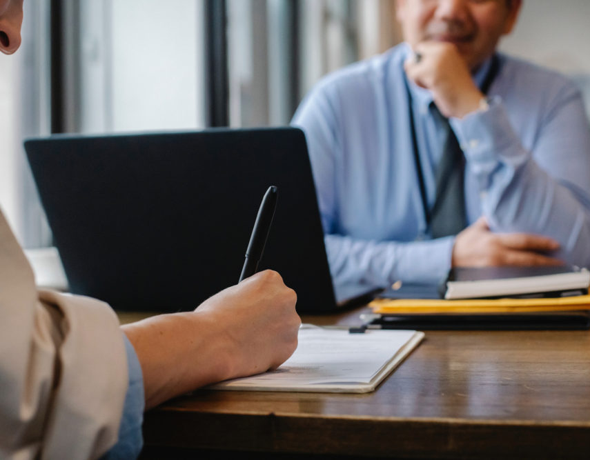 An over-the-shoulder view of a person writing with a pen on a clipboard at a wooden desk. Across from them, a man in a blue shirt and tie sits slightly out of focus, resting his chin on his hand. A laptop is open on the desk between them.