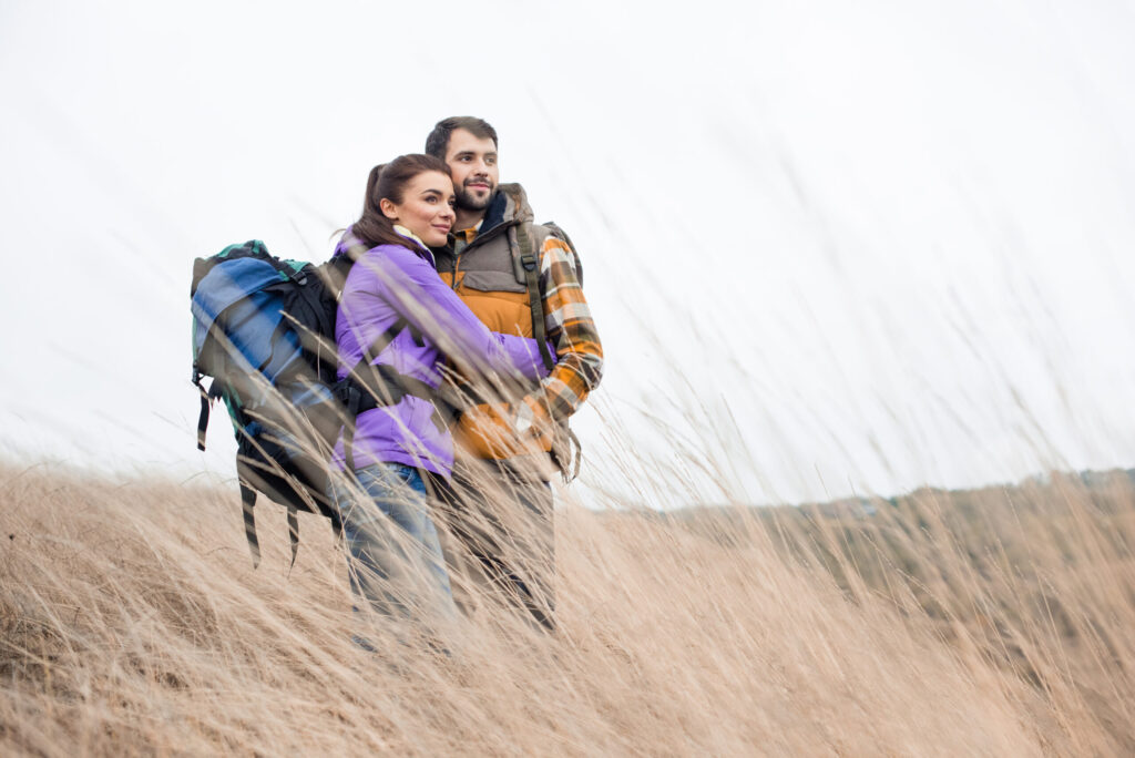 A young couple, both wearing large hiking backpacks and jackets, stand closely together in a field of tall, dry grass. The woman, in a purple jacket, wraps her arms around the man, who wears a plaid vest. They are both smiling and looking off into the distance to their right against an overcast sky.